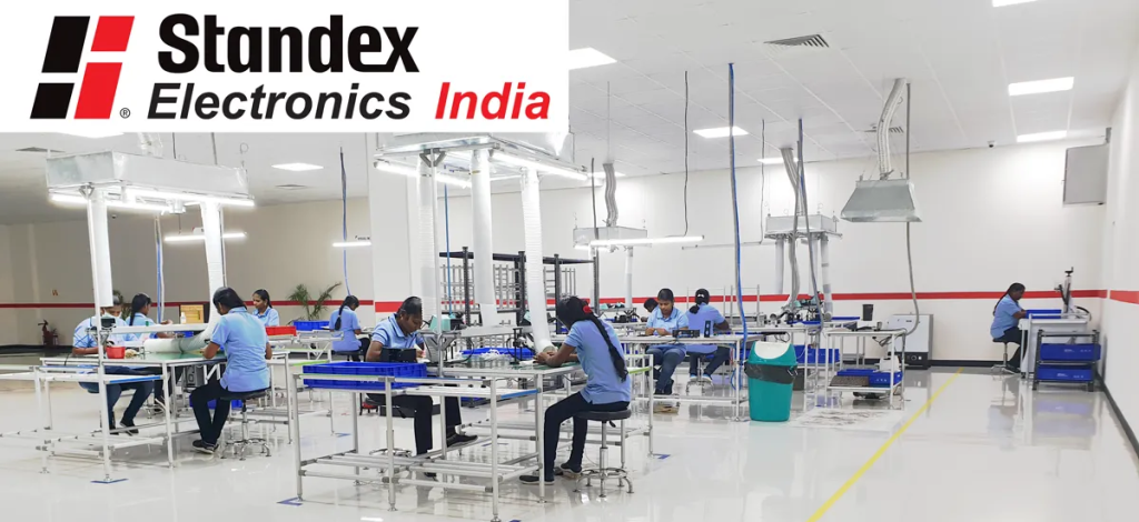 Workers in blue uniforms assemble electronic components at workstations in a bright, modern Standex India factory, highlighting ISO 9001:2015 certification and commitment to quality management. Overhead lights illuminate the workspace; logo appears top left. by Standex Detect